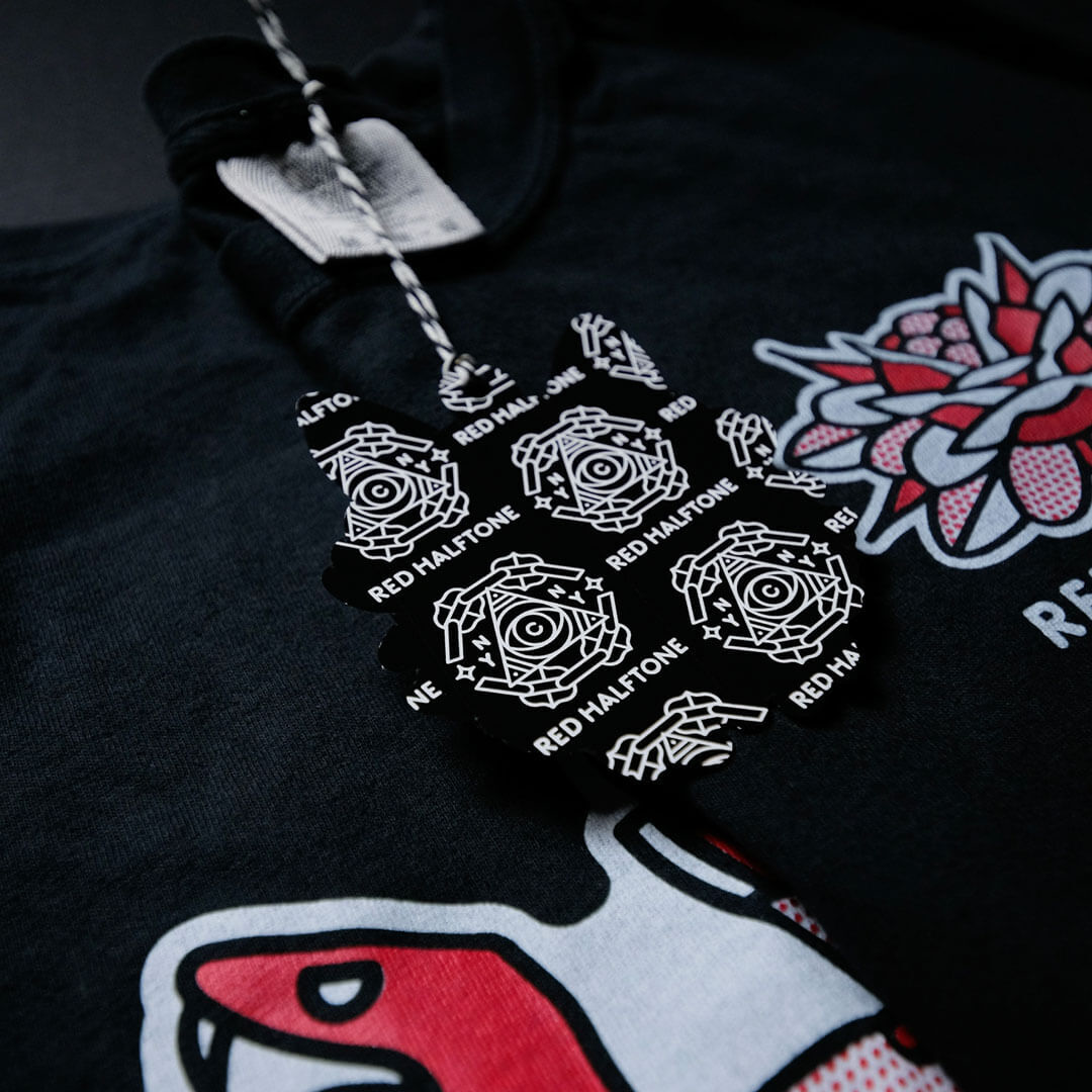 Schwarzes Shirt mit einem roten und weißen Grafikdesign, das ein florales Motiv zeigt. Ein schwarzes Etikett mit geometrischen Mustern und dem Text "RED HALFTONE" ist angebracht.