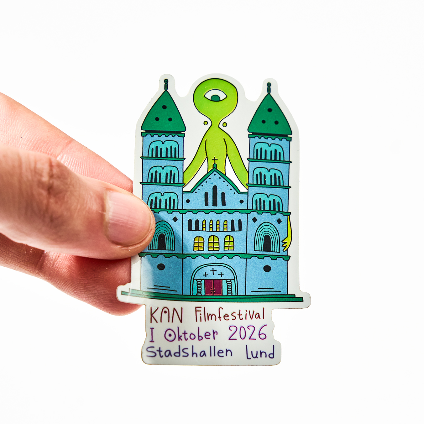Hand hält einen Freiform-Aufkleber für das KAN Filmfestival, Oktober 2026 in der Stadshallen Lund, mit einer Kathedrale und einem grünen einäugigen Alien