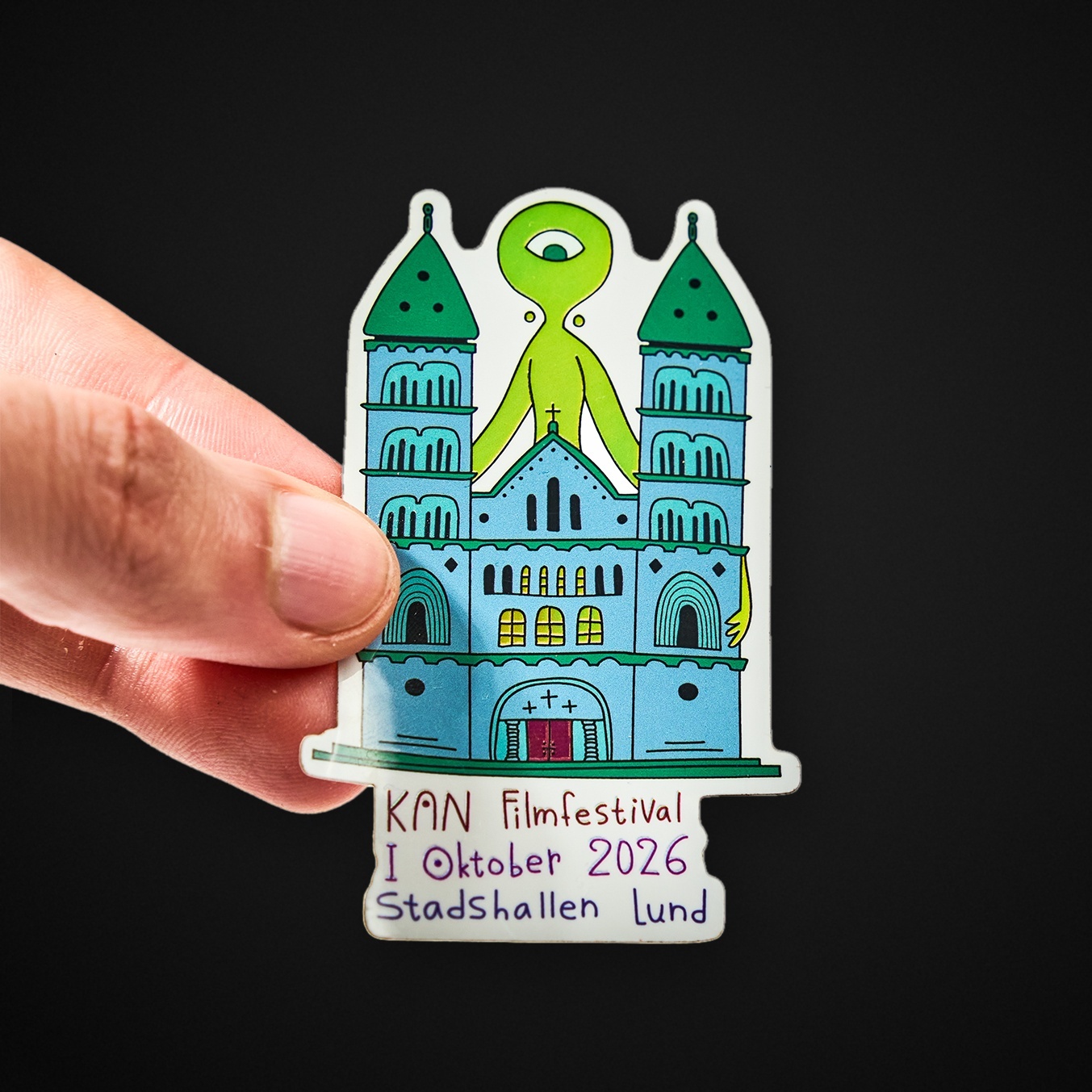 Hand hält einen Freiform-Sticker mit einer Kathedrale und einem grünen einäugigen Wesen, Aufschrift „KAN Filmfestival I Oktober 2026 Stadshallen Lund“