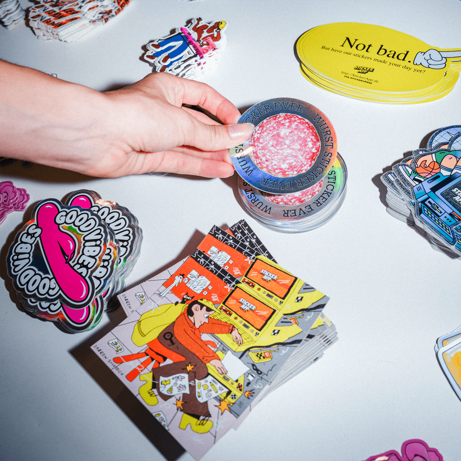 Hand hält einen runden holografischen Salami-Sticker über einem Tisch mit Freiform-Stickern in verschiedenen Finishes