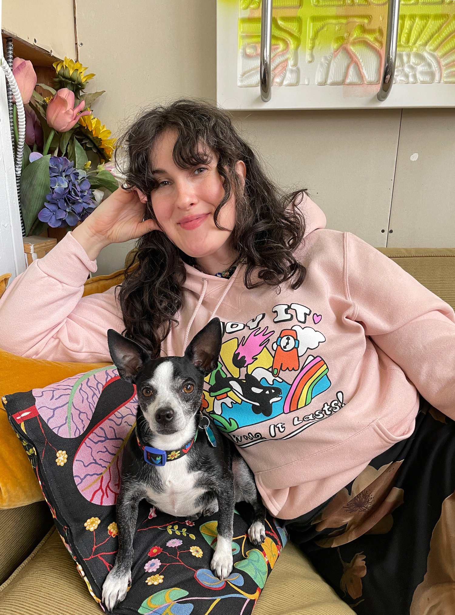 Lächelnde Grace Miceli im bunten Hoodie neben ihrem Hund, der auf einem Kissen liegt